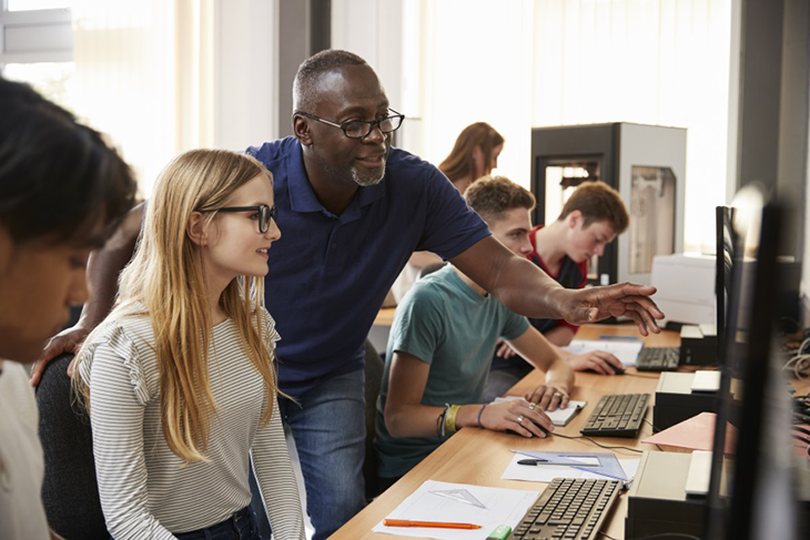 Profesor señala algo en el monitor a una alumna