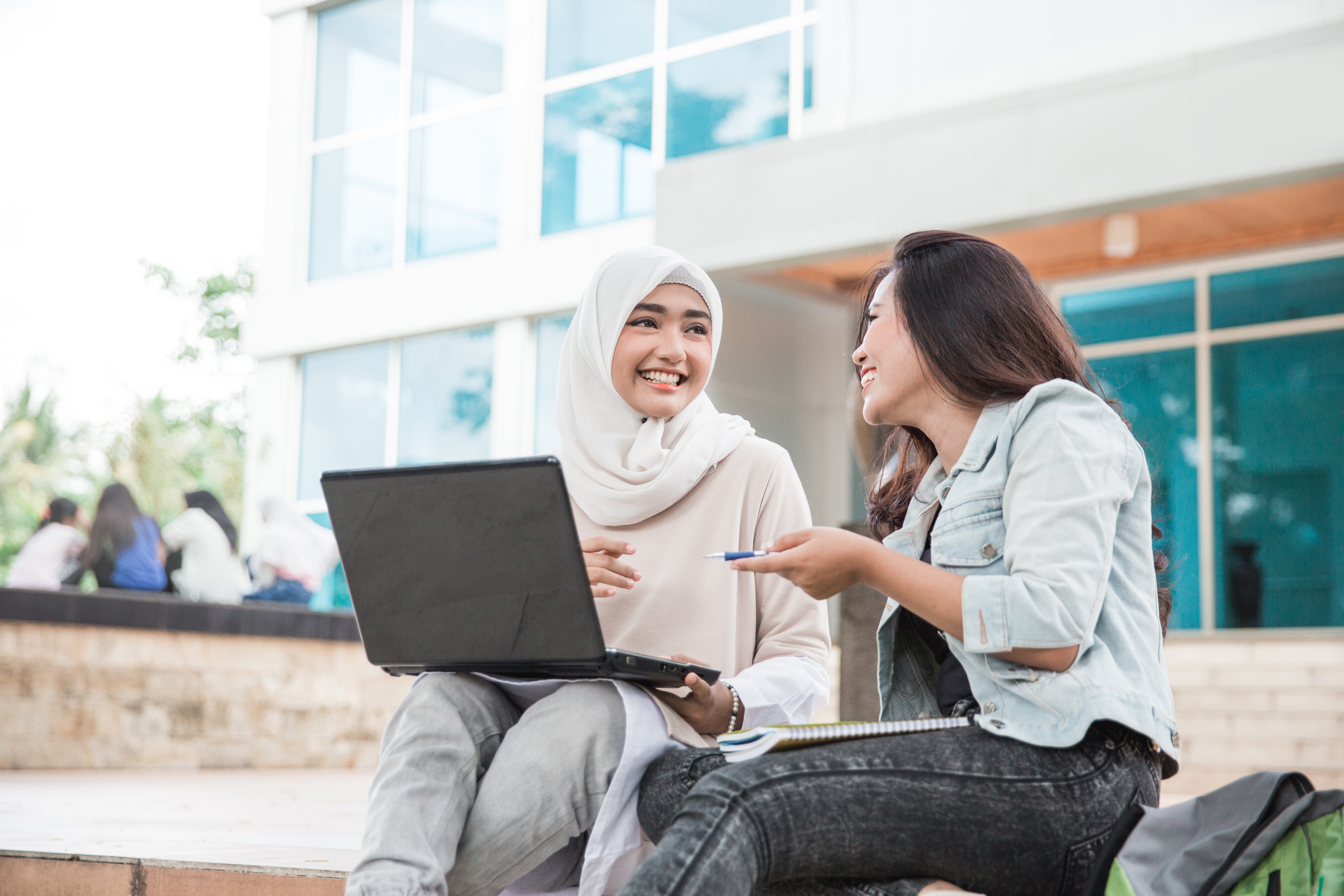 A young woman wearing a headscarf works on the computer and smiles with an adult woman as they sit on stairs outside.