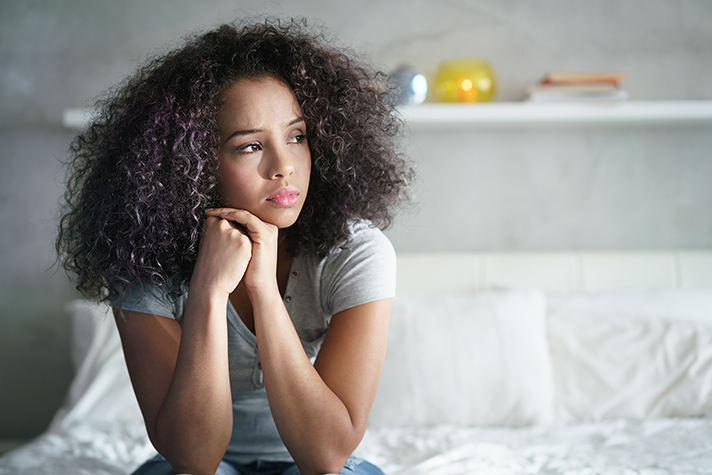A teenage girl sits on the edge of a bed resting her head in her hands as she looks off. Her expression conveys worry, sadness, and thoughtfulness.