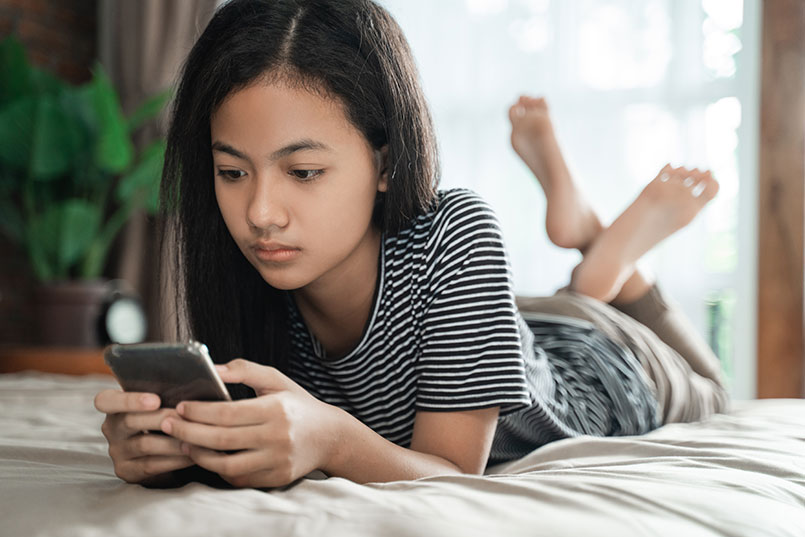 침대 위에 편히 엎드려 휴대전화를 하는 십대 소녀