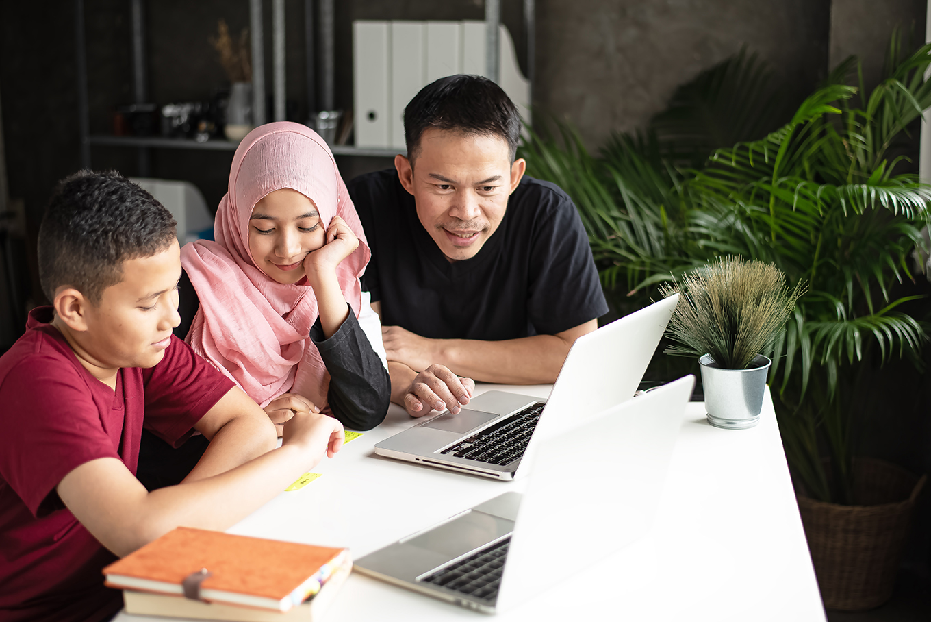 A man, a girl, and a boy are sitting at a table and looking at two laptops.