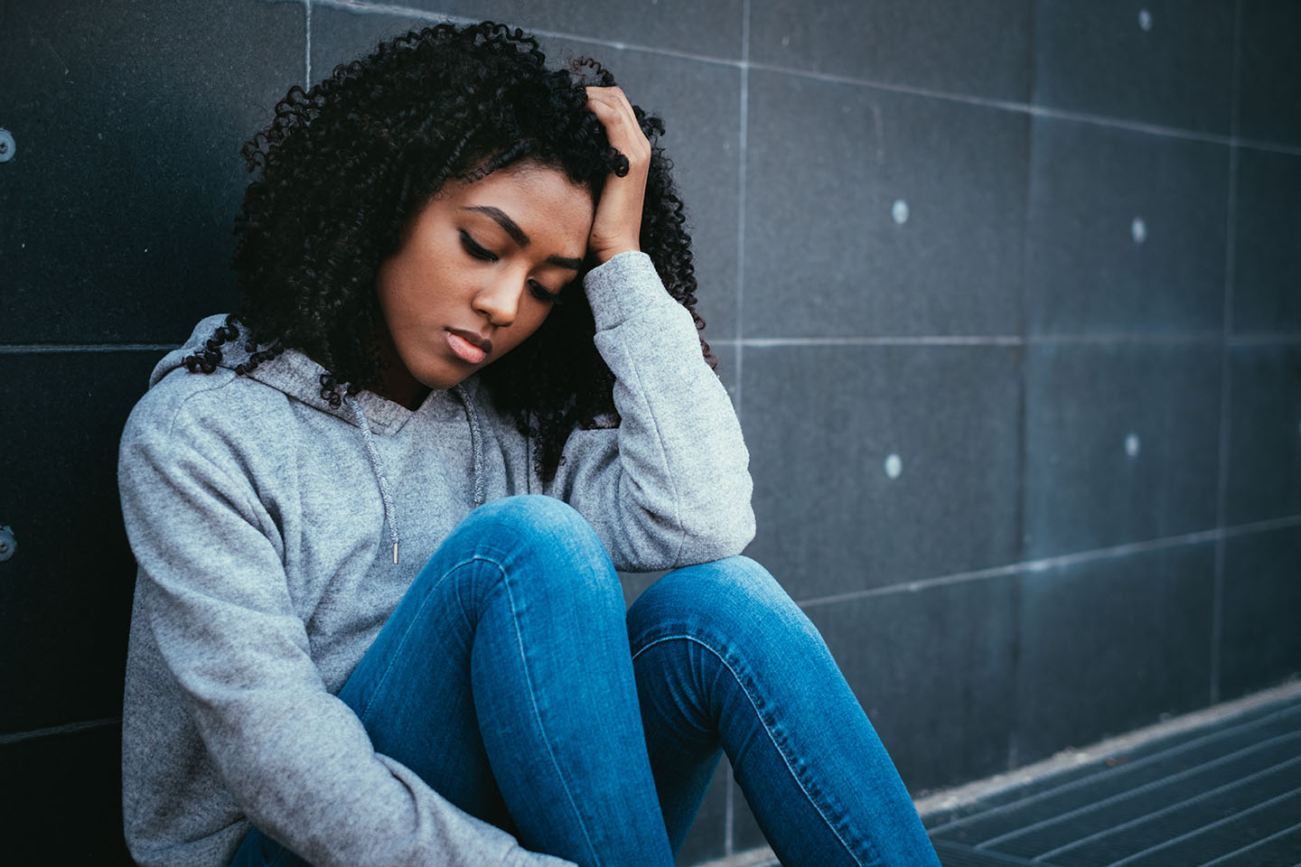 A teen girl looks sad as she sits on the ground against a wall and rests her head in her hand.