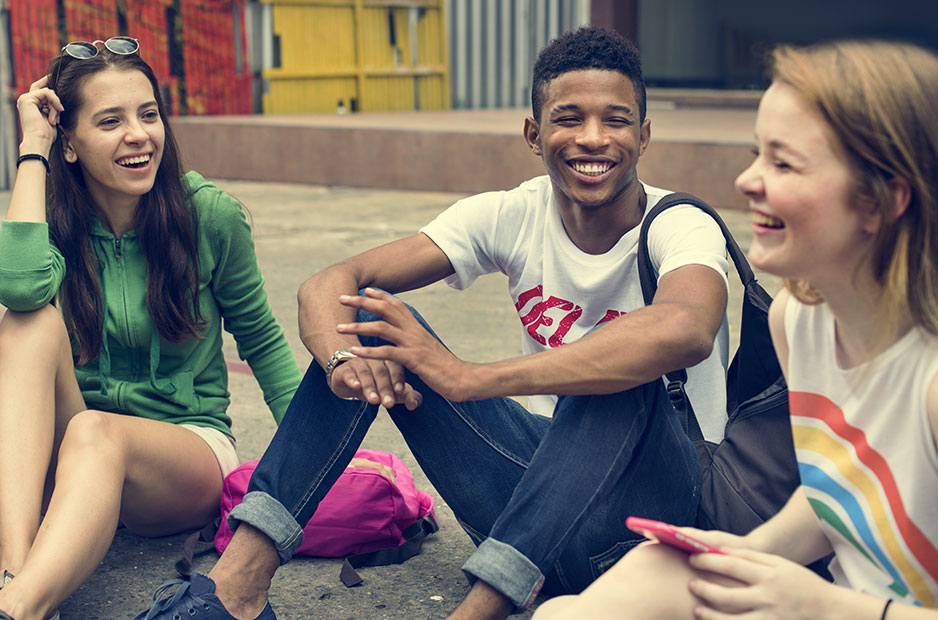 A group of teenage friends sit on the ground smiling and laughing together.