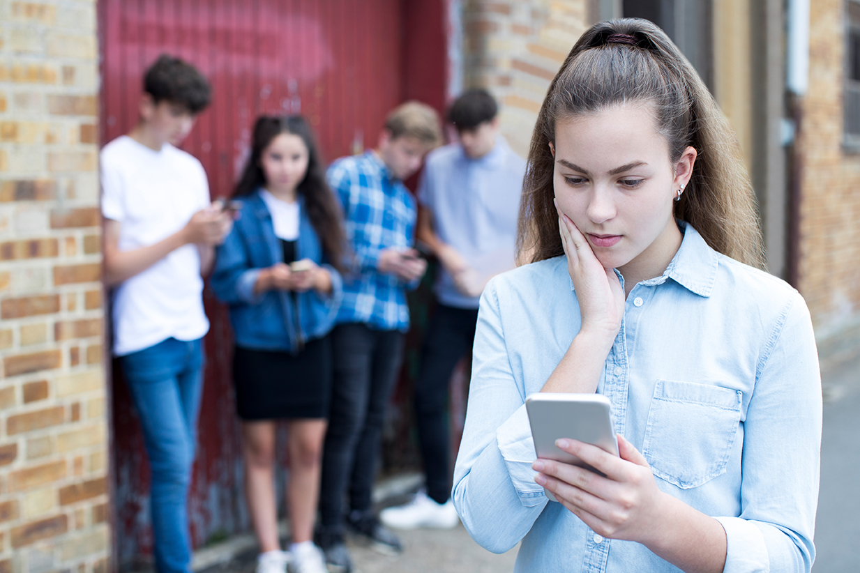 휴대폰을 심각하게 쳐다보고 있는 십대 소녀 그리고 뒤에서 모여 휴대폰을 보고 있는 다른 십대들.