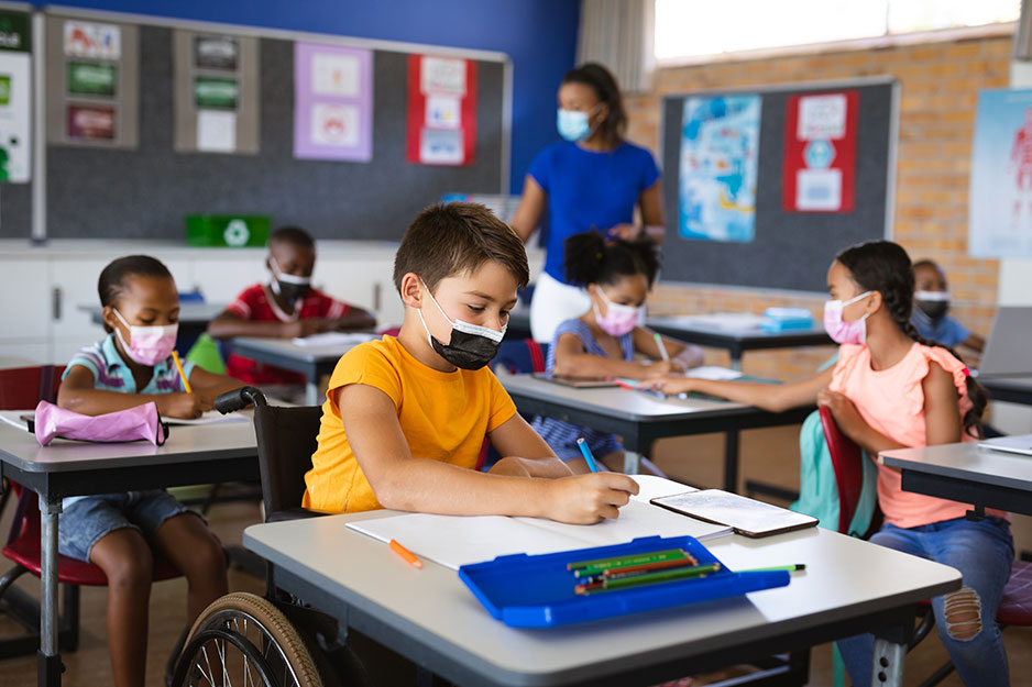 휠체어를 탄 한 아이가 다른 아이들과 선생님과 함께 모두 마스크를 쓰고 있는 교실에서 책상에 앉아 있습니다.
