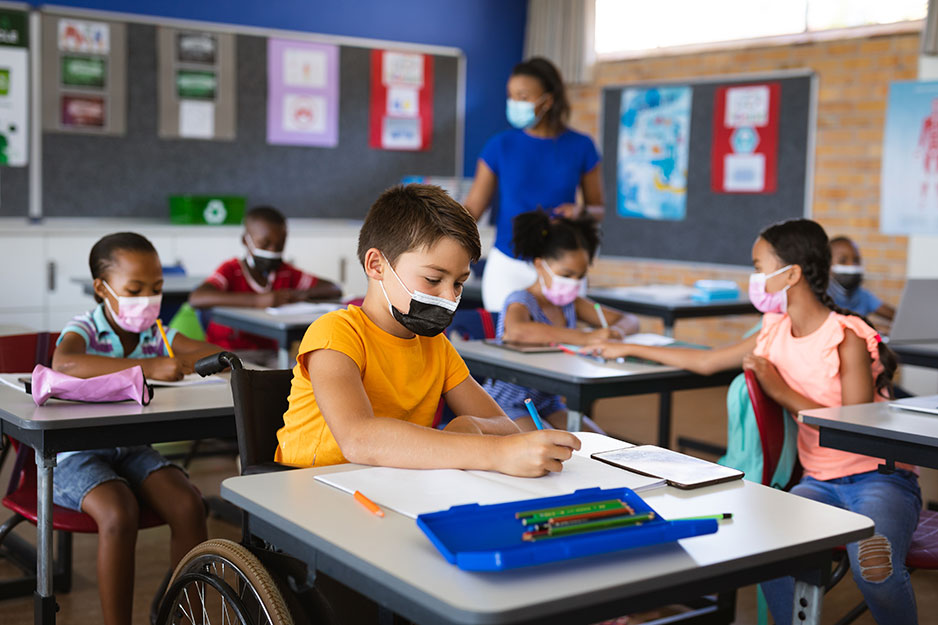 A child in a wheelchair sits at a desk in a classroom with other children and a teacher all wearing masks.