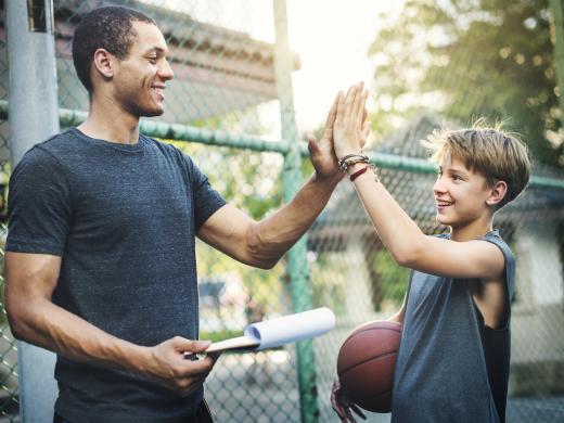 A young boy carrying a basketball is high-fiving an adult male carrying a clipboard on an outdoor basketball court.