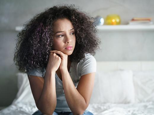 Una adolescente sentada en el borde de la cama, con la cabeza apoyada en sus manos, luciendo apagada. Su expresión demuestra preocupación, tristeza y reflexión.