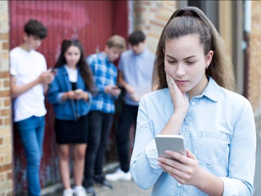 A teen girl looks at her cellphone with concern, as other teens gather and look at their cellphones in the background.
